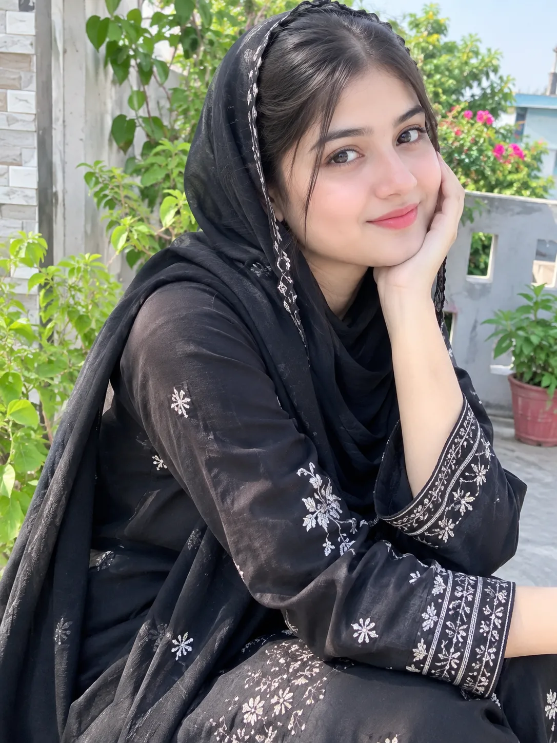 : A beautiful young South Asian woman wearing an elegant black embroidered outfit and hijab, sitting gracefully on a green terrace garden,