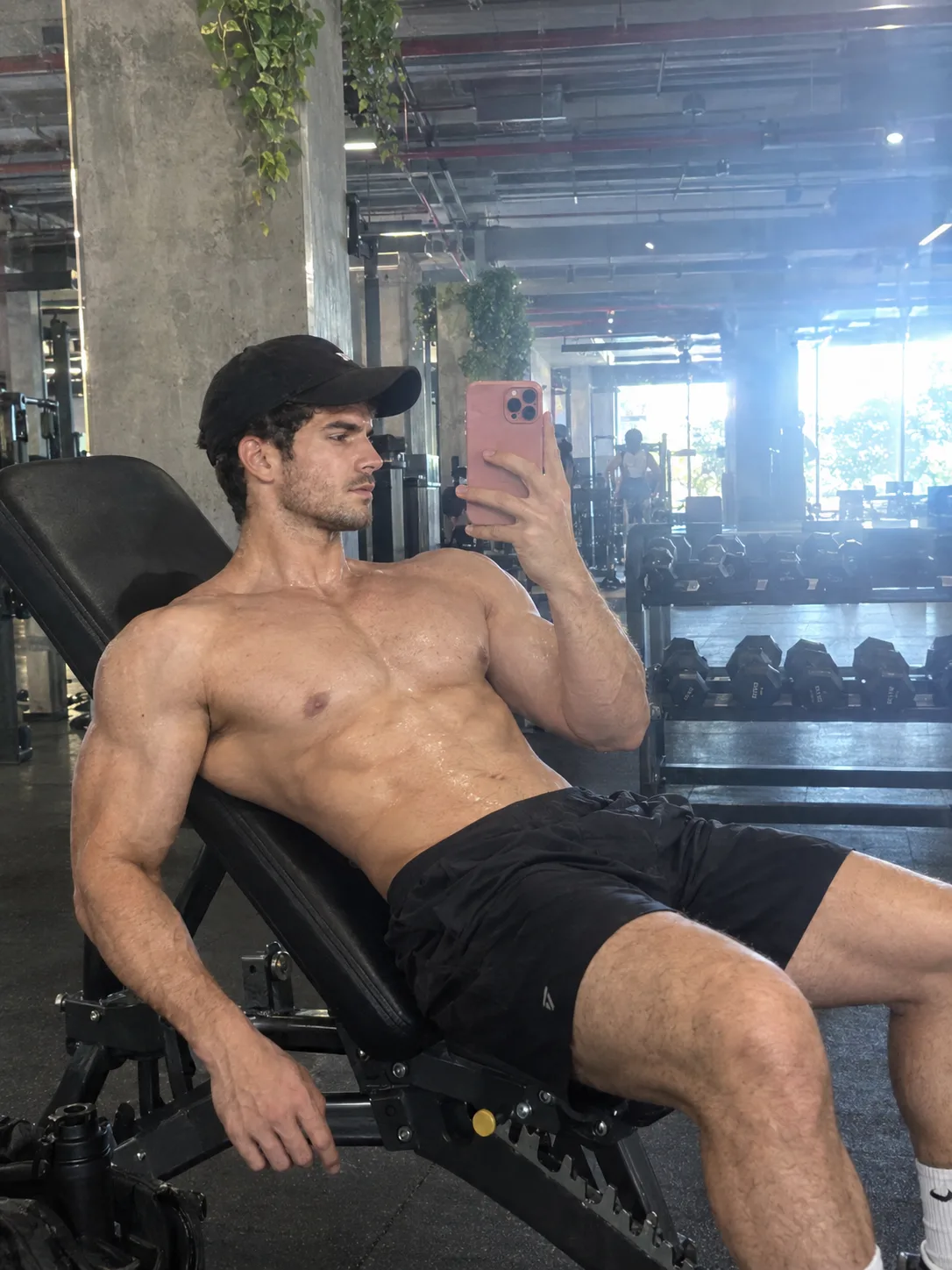 Vertical iPhone 17 Pro Max mirror-adjacent selfie, shot from a low reclined angle, subject leaning back on an inclined black gym bench