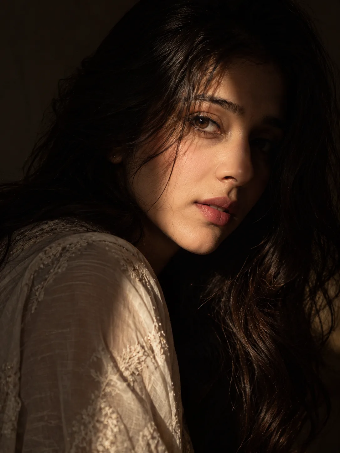 Create A cinematic close-up portrait of a young woman with long, dark, wavy hair and expressive brown eyes, captured in dramatic low-key