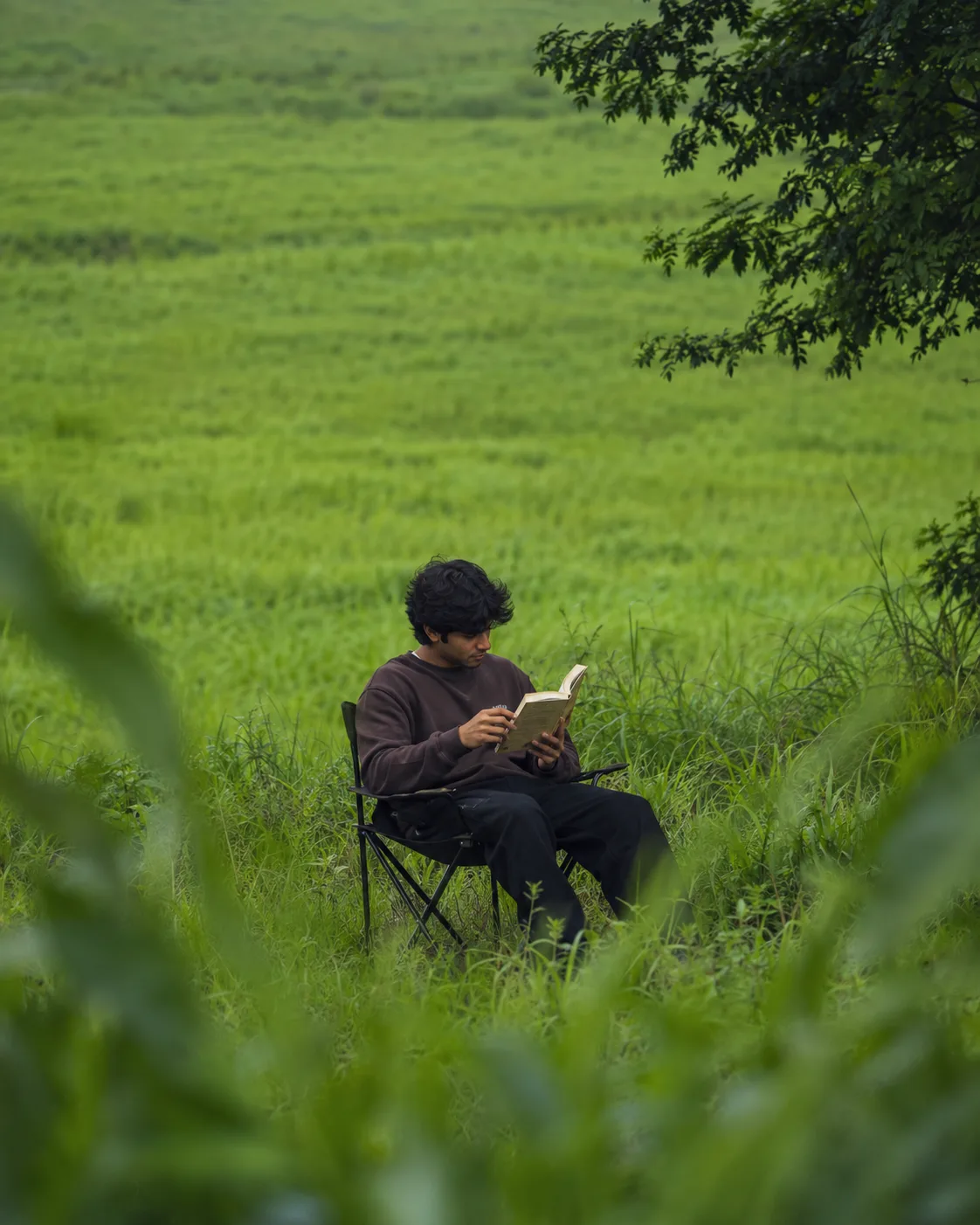Create an ultra-realistic cinematic lifestyle photograph of a young South Indian man sitting alone in the middle of a vast lush green