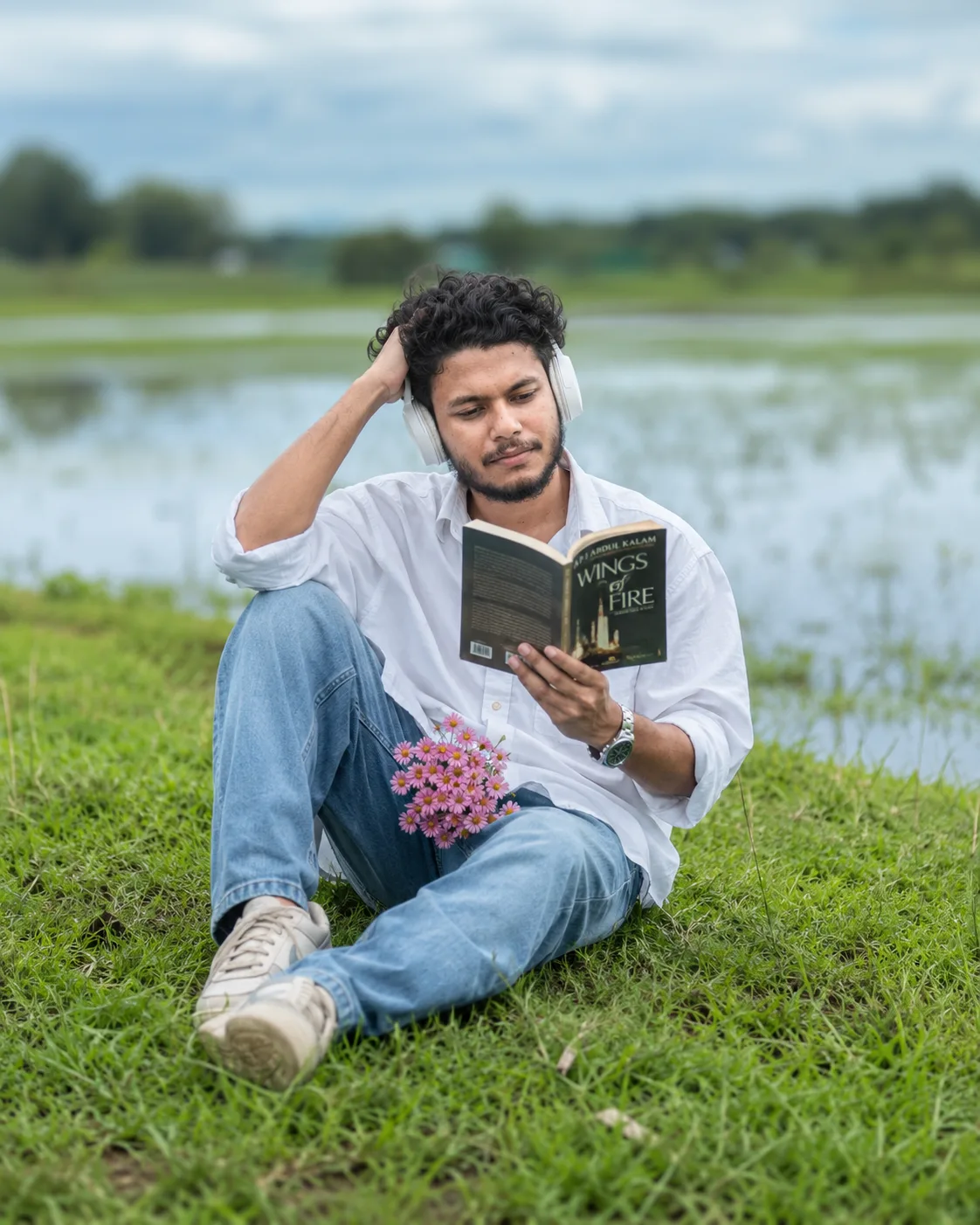 A young Indian man with curly black hair and a short beard is sitting casually on lush green grass beside a calm lake.
