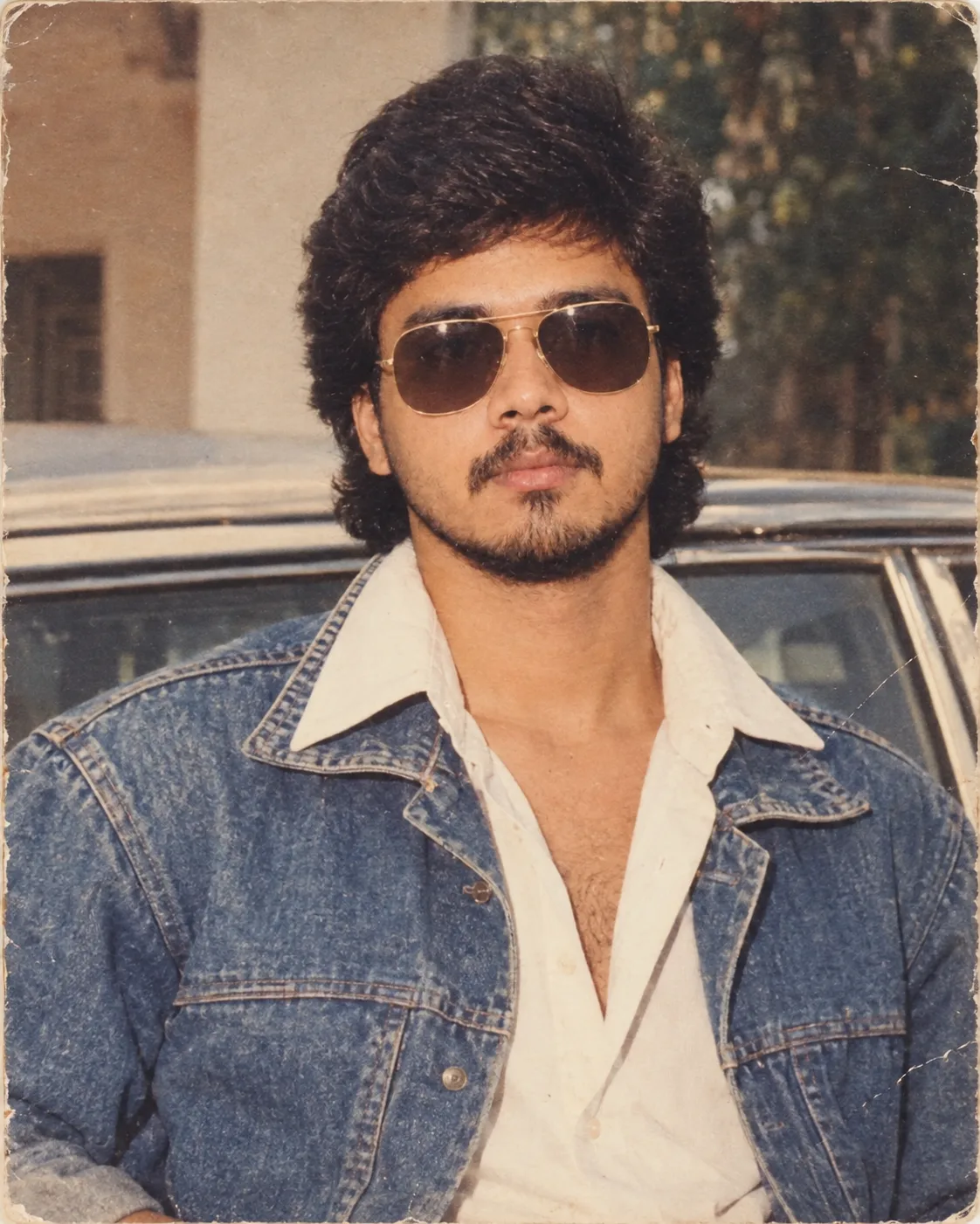 Create an ultra-realistic vintage 1980s Indian cinematic portrait of a young man standing confidently in front of a classic vintage car.