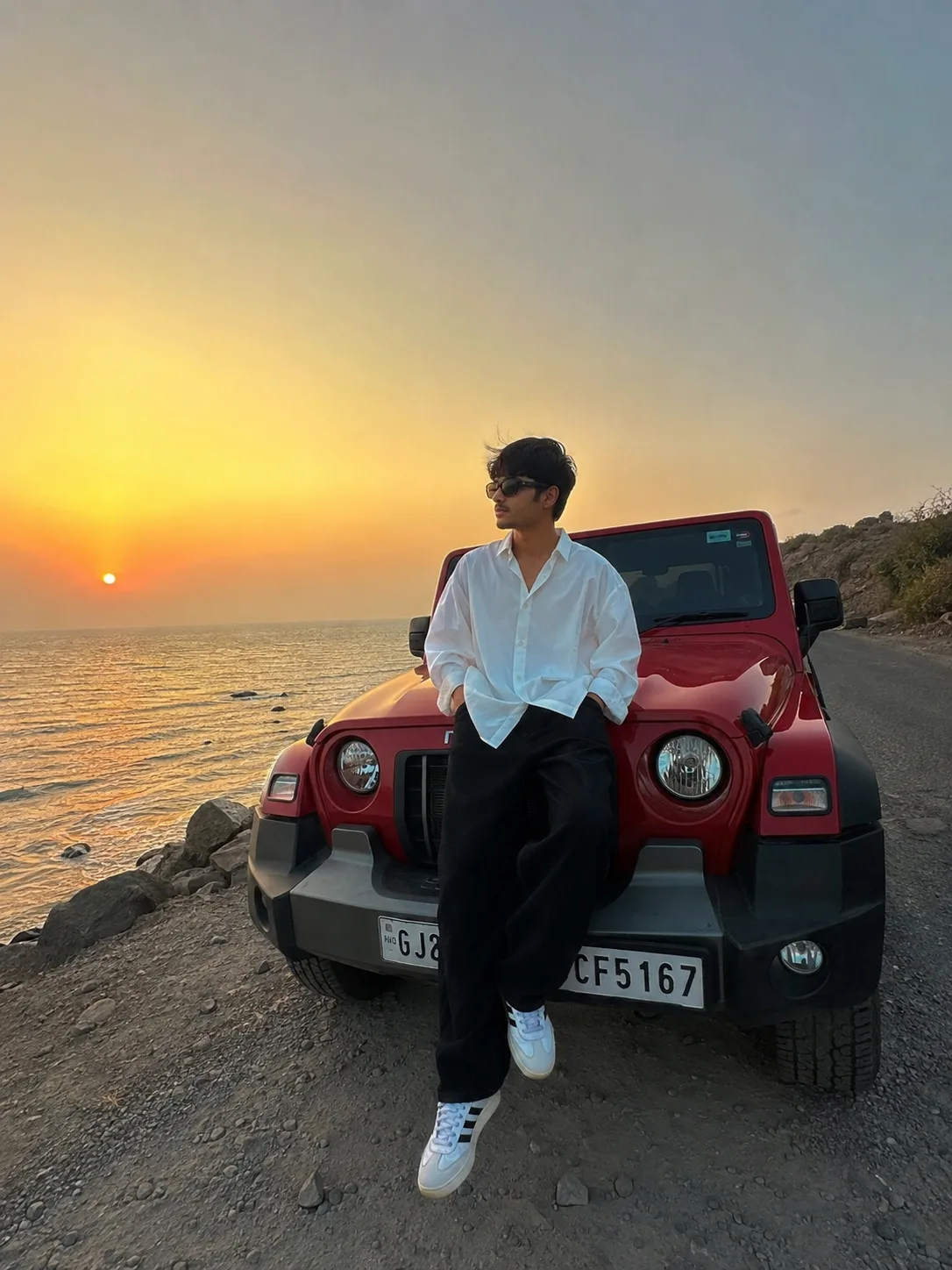 An ultra-realistic casual smartphone photograph in 3:4 ratio — a young man sitting casually on the hood of a red Mahindra Thar parked