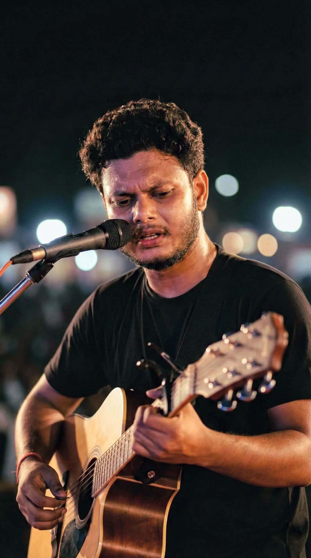 A young Indian man in his late 20s with short curly black hair, light stubble beard and mustache, singing passionately into a black