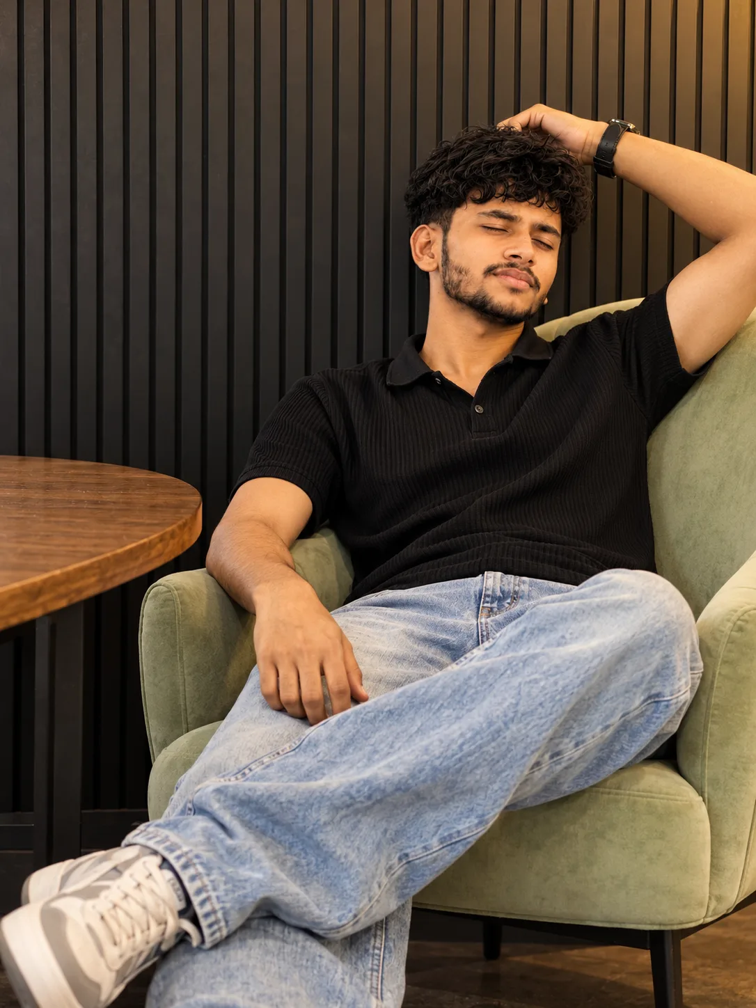 Ultra-realistic professional DSLR photograph of a slim young South Asian man reclining casually in a pale sage-green upholstered lounge