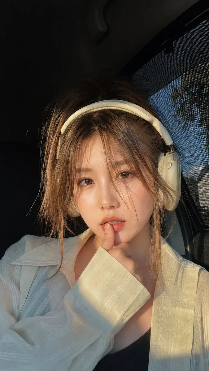 Ultra-realistic close-up portrait of a beautiful young woman sitting inside a car during golden hour, wearing elegant cream-white over-ear