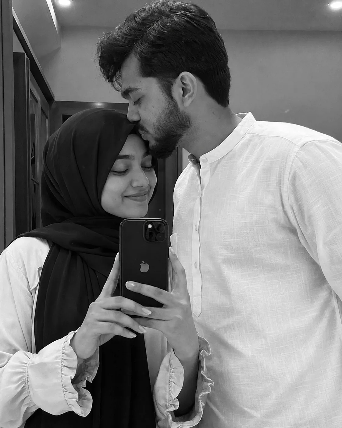 A realistic, intimate black and white candid mirror selfie of a young South Asian couple.