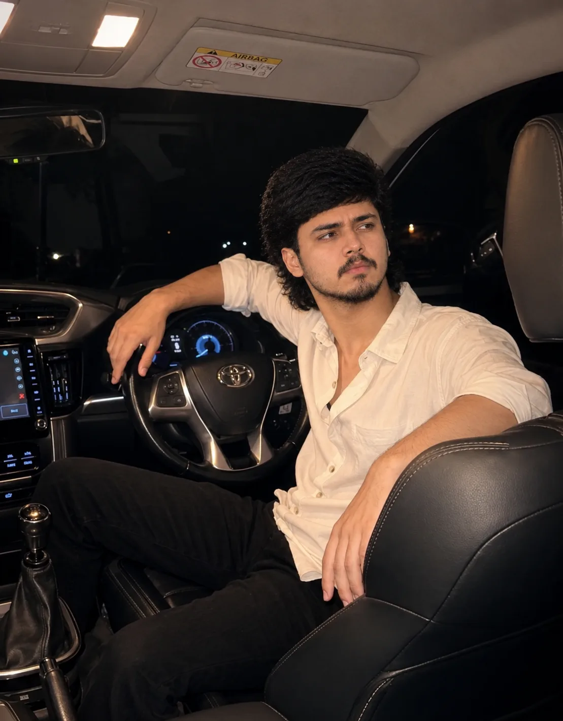 A photorealistic cinematic nighttime portrait of a young South Asian man sitting casually in the driver’s seat of a modern Toyota car.