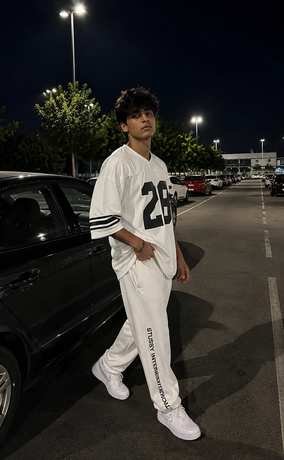 Ultra-realistic natural smartphone night photography of a young man as reference image standing in an outdoor parking lot beside a car.