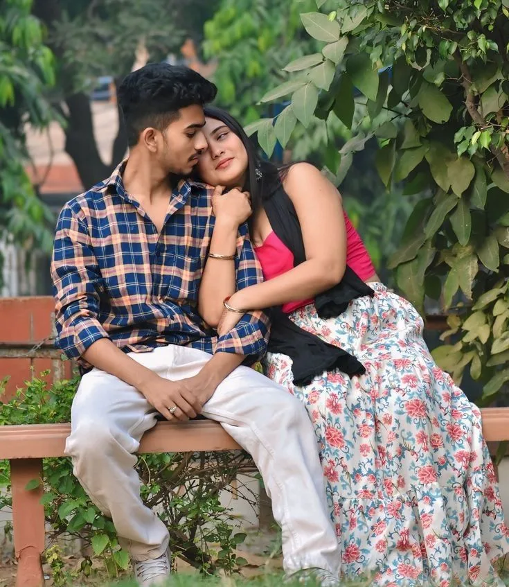 A photorealistic vertical shot of the young South Asian couple seated outdoors on a low park bench, faithfully preserving both subjects'