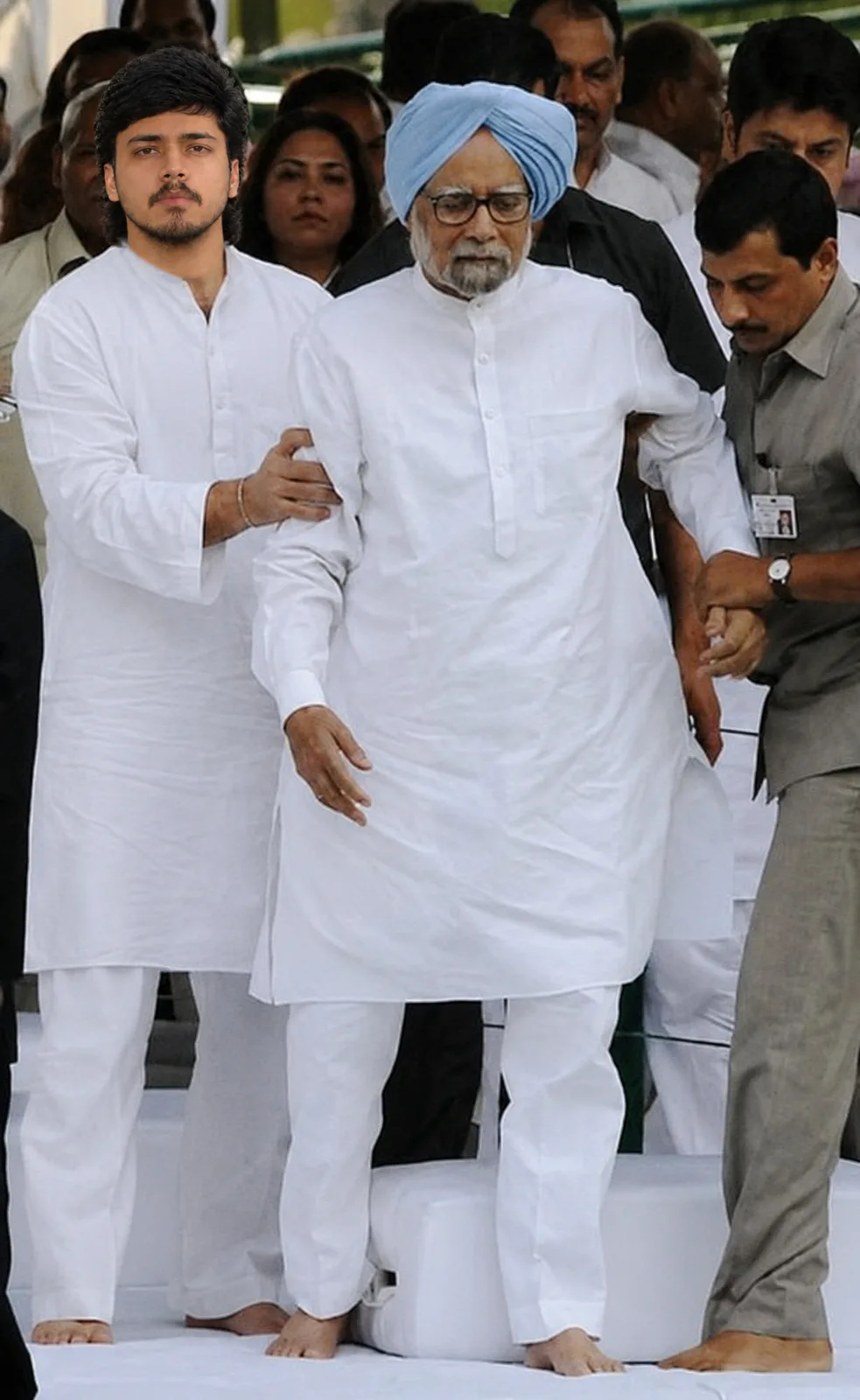 A realistic documentary-style photograph of a young Indian man walking respectfully beside an elderly Indian statesman wearing a crisp