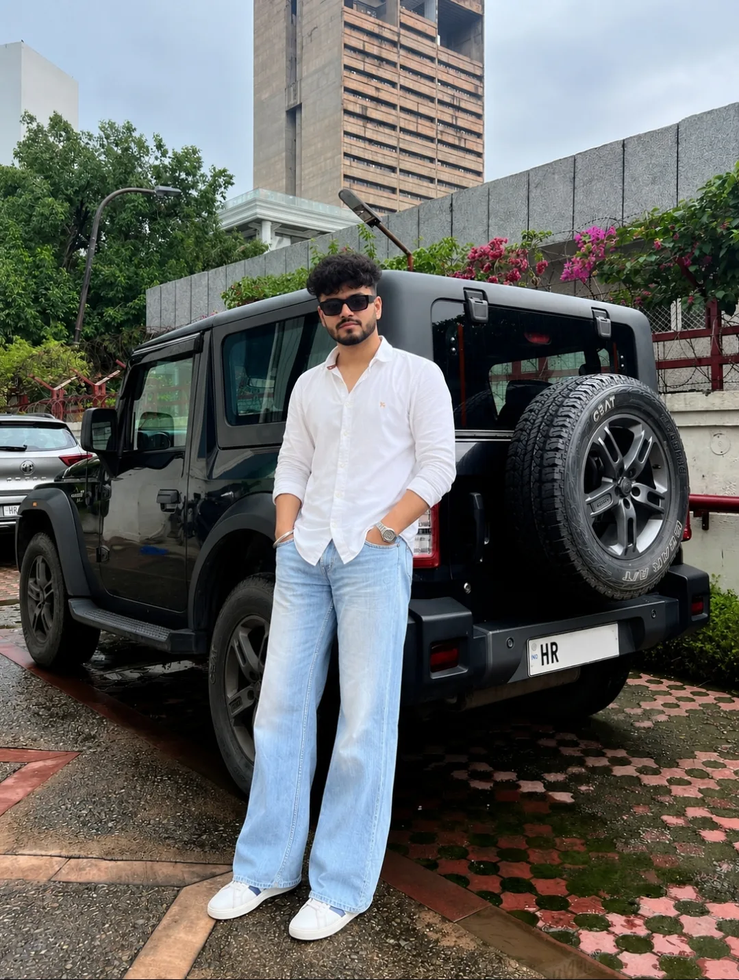 Create an ultra-realistic DSLR photograph of the same young man standing casually beside the rear side of a black Mahindra Thar, preserving