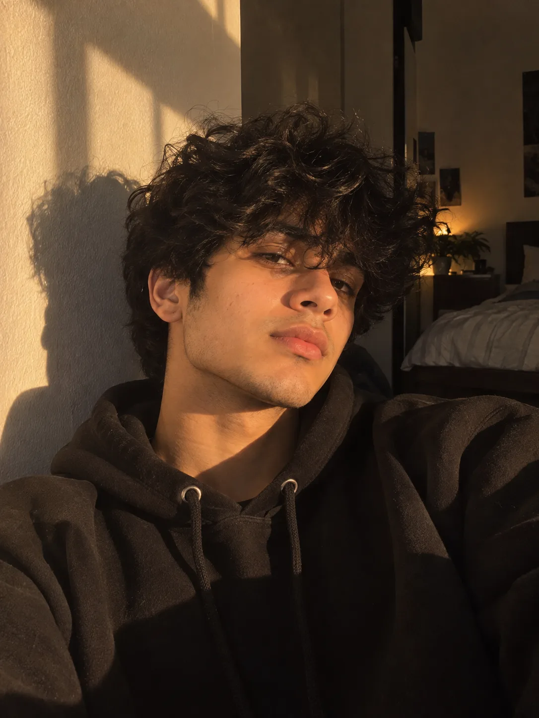 A cinematic close-up portrait of a stylish young man relaxing indoors beside a sunlit wall, captured in an intimate candid selfie