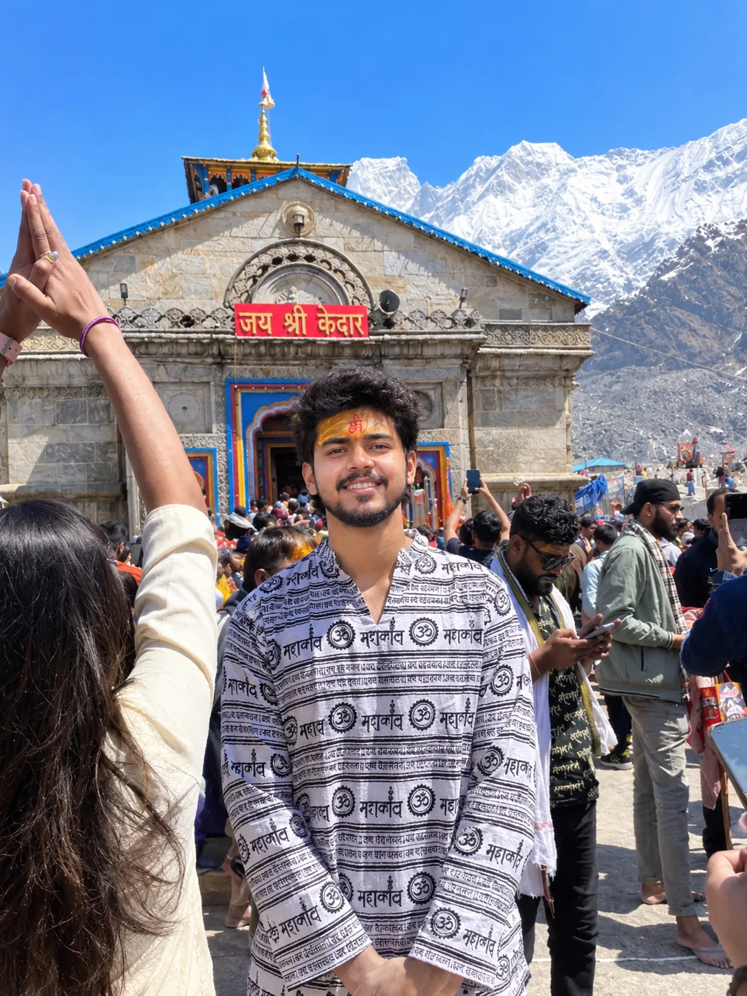 A photorealistic cinematic travel portrait of a young Indian man standing in front of the sacred Kedarnath Temple in the Himalayas.
