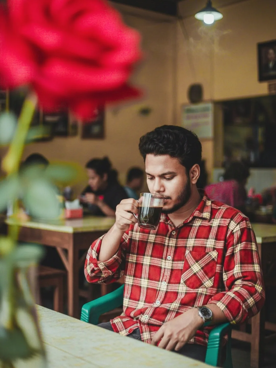 A young man with dark, wavy hair and a beard, wearing a red and beige plaid shirt, is sitting at a table in a cozy cafe or restaurant.