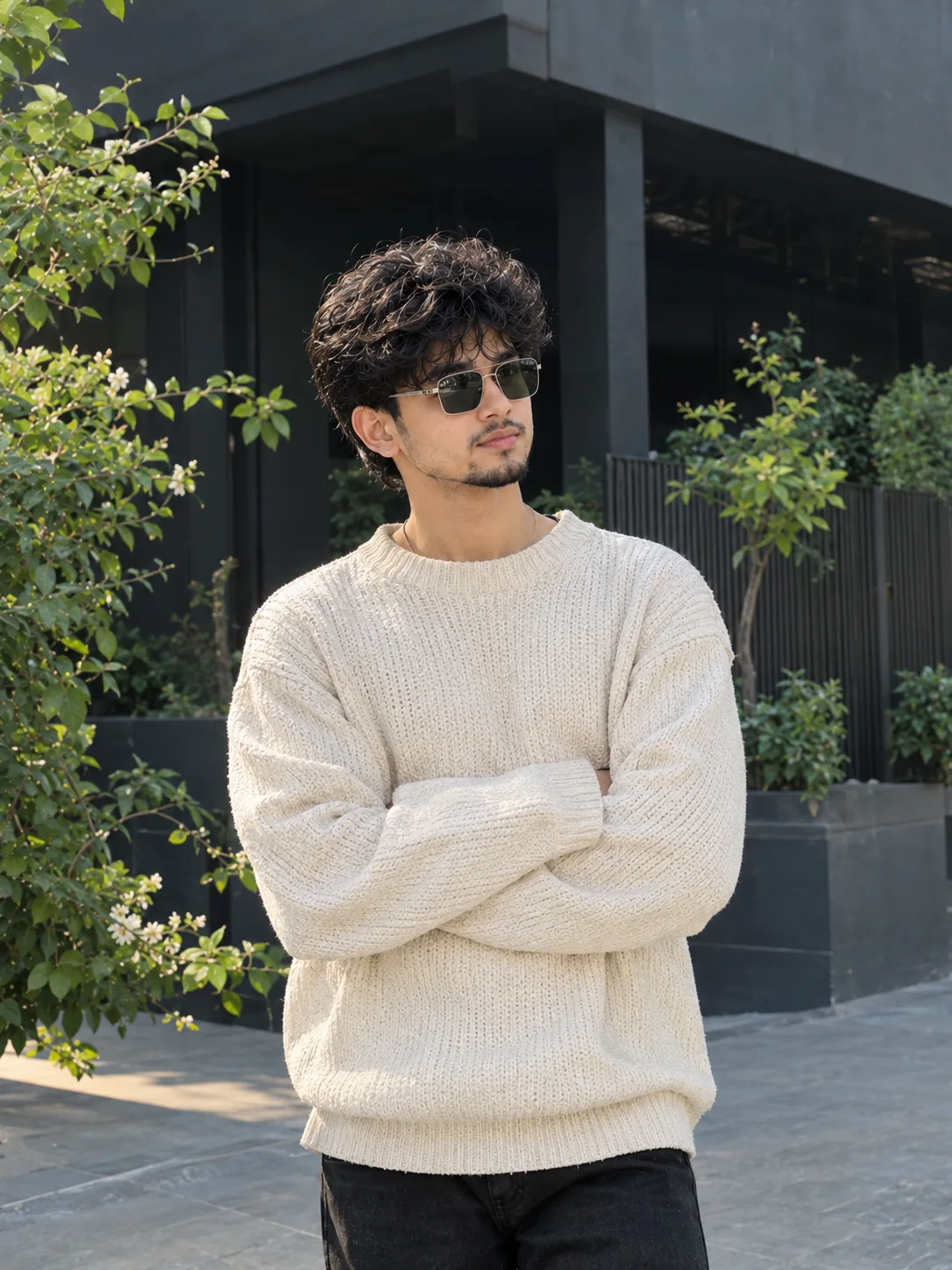 Ultra realistic photograph of a handsome young Indian man posing casually in a modern outdoor setting, wearing a textured oversized