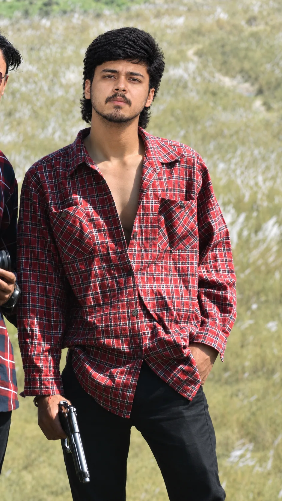 Create an ultra-realistic cinematic vertical portrait of a young South Asian man standing confidently outdoors in a sunlit grassy field.