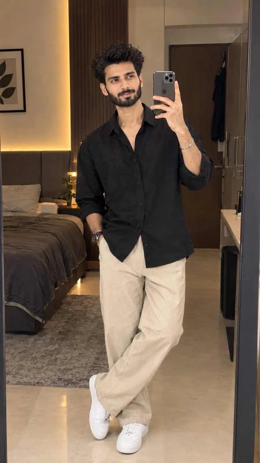 Ultra-realistic 9:16 mirror selfie of a stylish 20-year-old Indian man standing confidently in a modern bedroom, holding an iPhone in one
