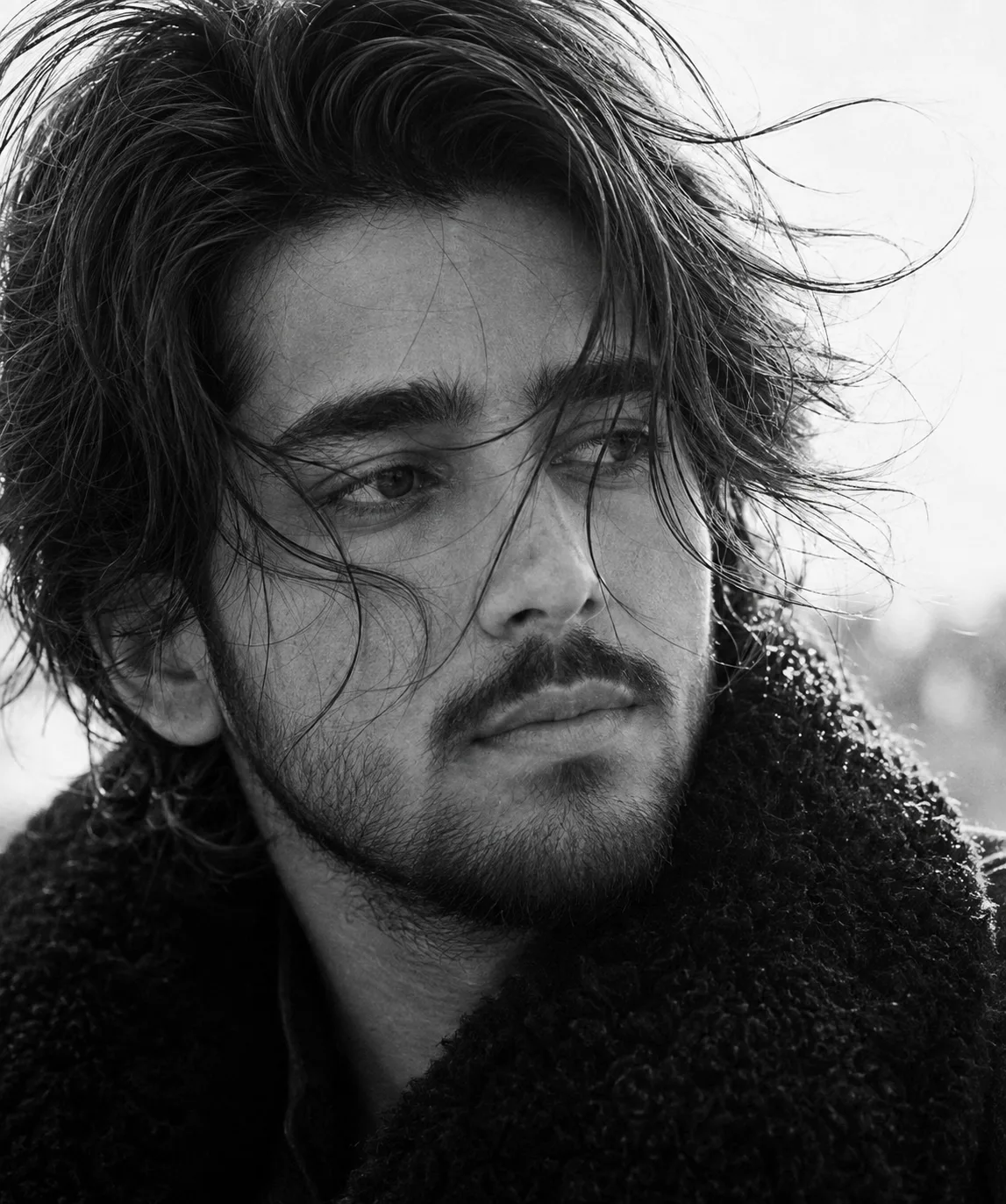 A highly detailed, photorealistic black-and-white close-up portrait of a young adult man photographed outdoors in a cold, windy