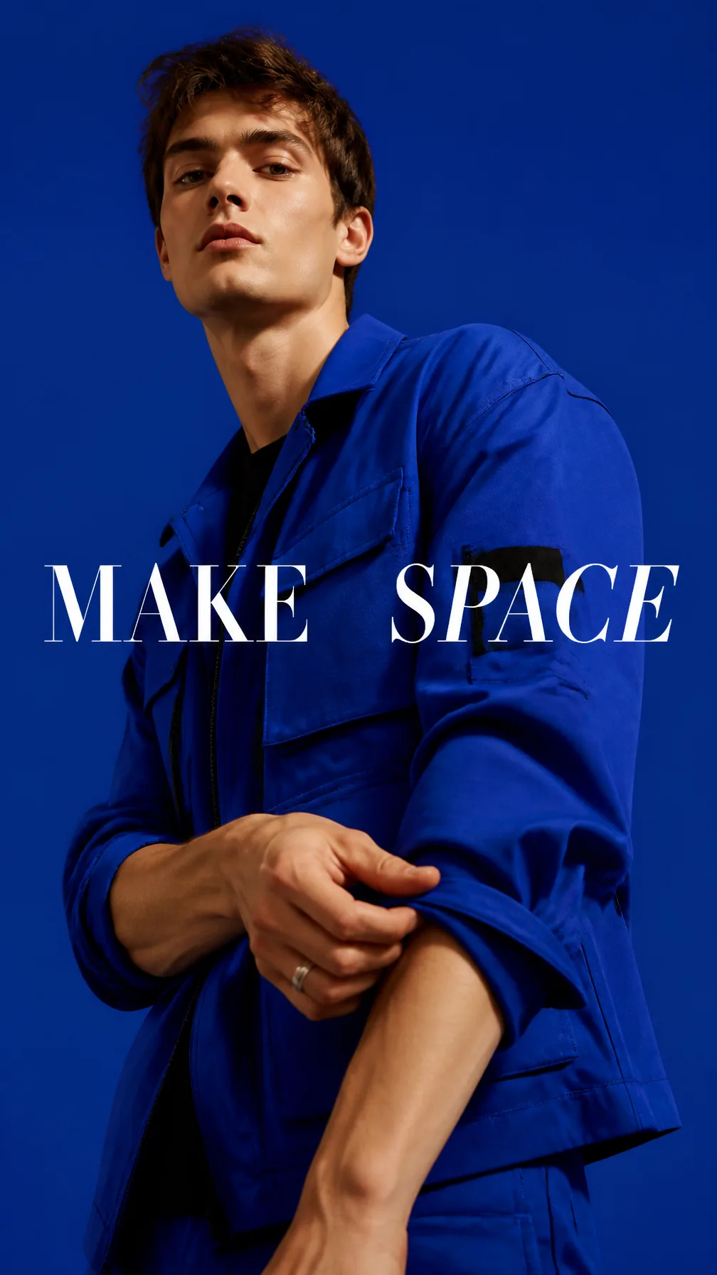 Premium editorial campaign.Main Subject: Young male model wearing a vivid cobalt utility jacket.Pose: Slowly rolling down one shirt sleeve