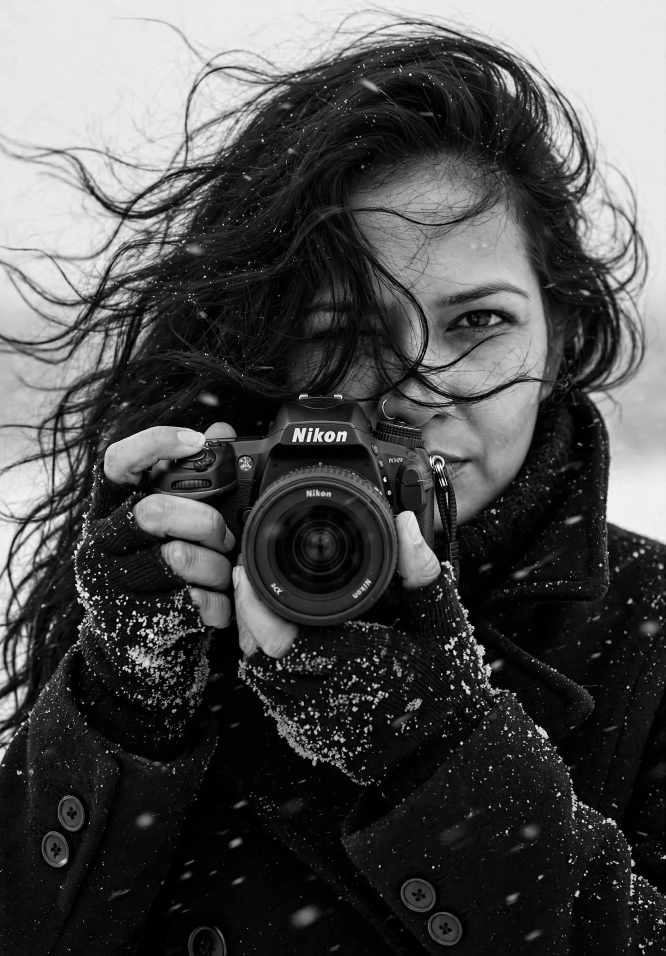 Create a black and white portrait exactly like reference: SUBJECT: Woman photographer holding Nikon DSLR camera in snow, hair blowing in