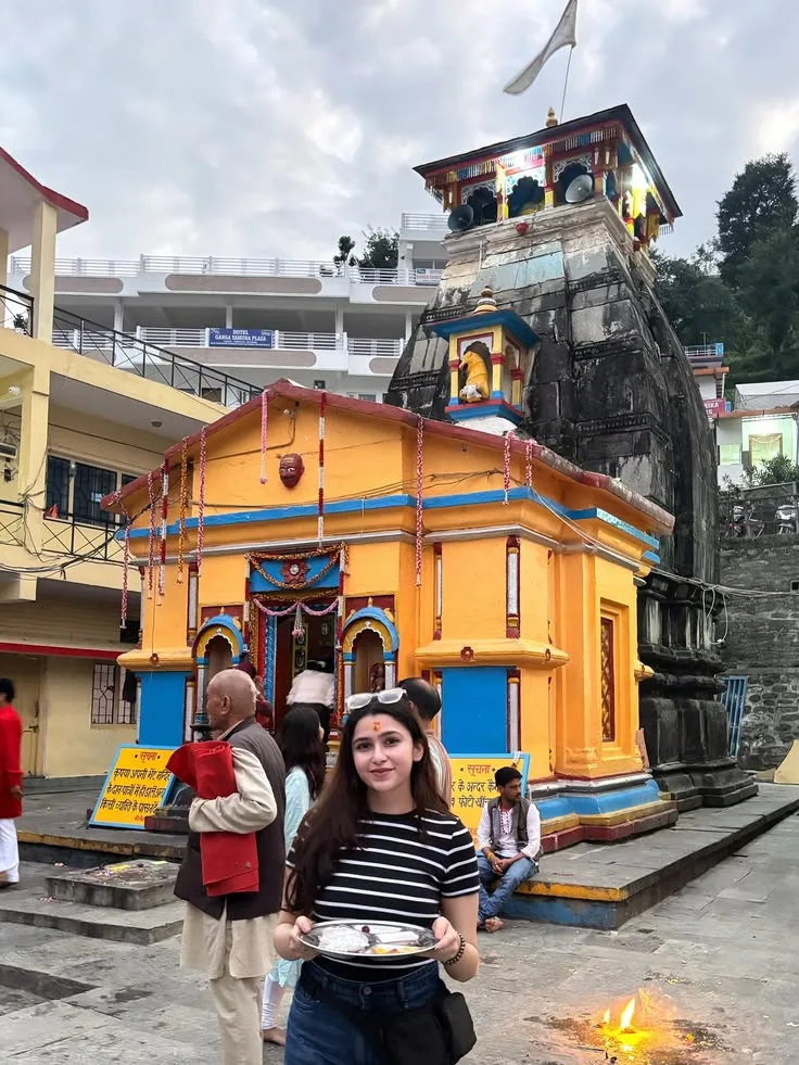 Create a photorealistic vertical travel photograph of a young Indian woman standing in front of a colorful traditional Hindu temple in the