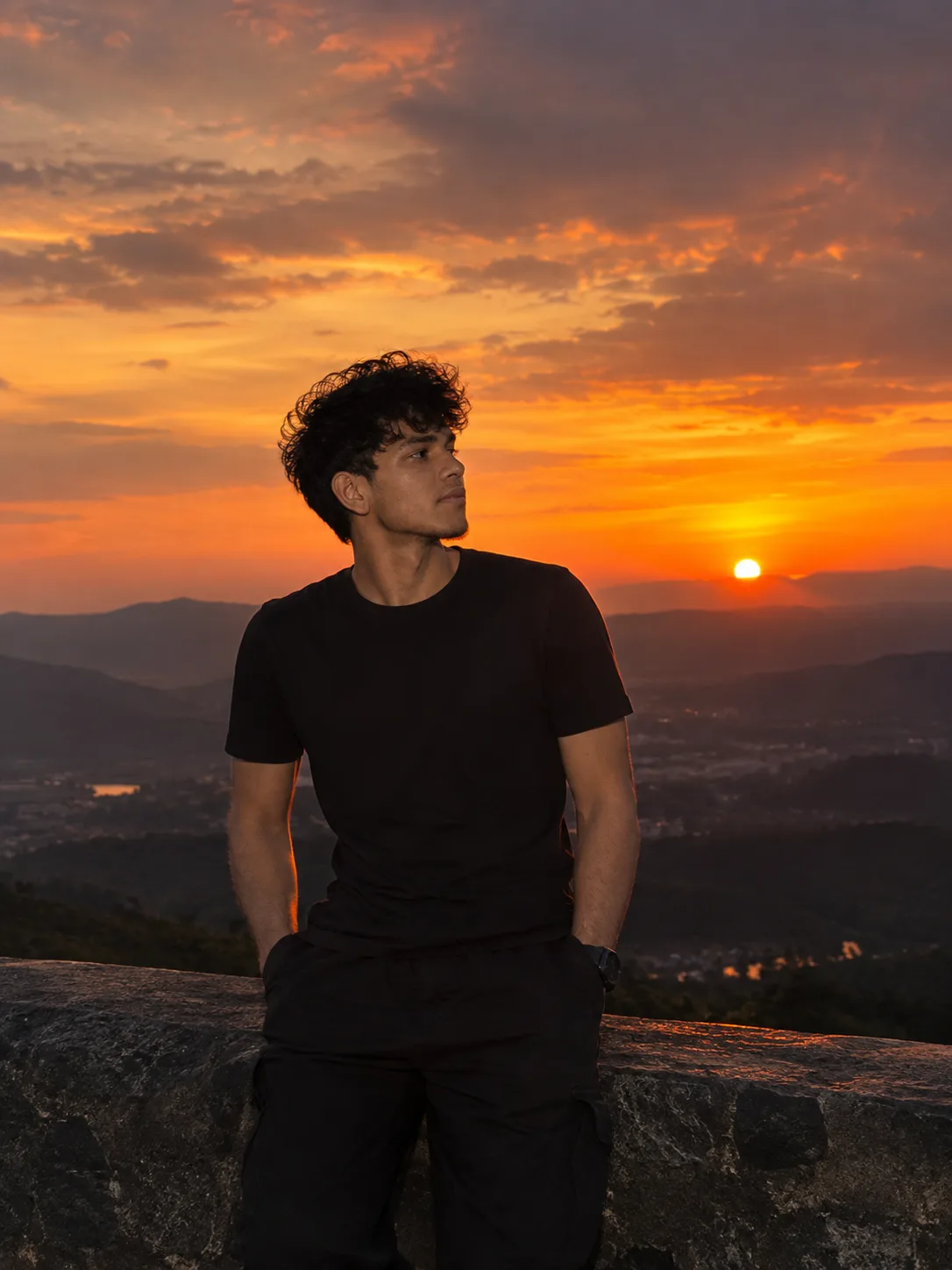 Young man standing at a mountain viewpoint during a stunning sunset, golden-orange sky, dramatic clouds, overlooking valleys and hills,