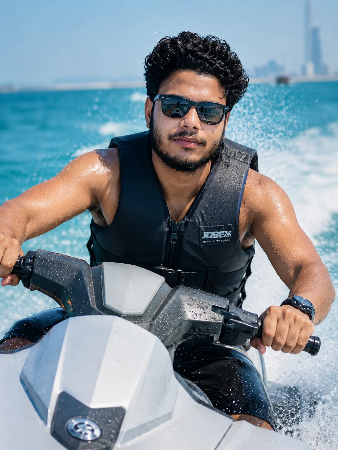 A dynamic close-up action shot of a young Indian man with curly black hair and a short beard, wearing black sunglasses, riding a white and