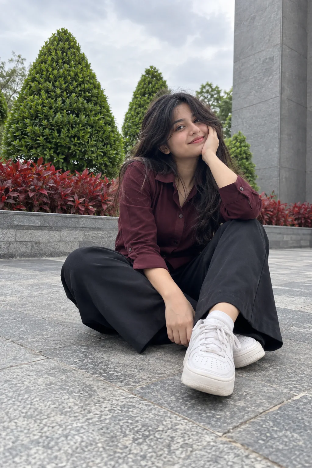 A candid, low-angle full-body photograph of a young Indian woman sitting comfortably directly on a grey stone paved floor outdoors.