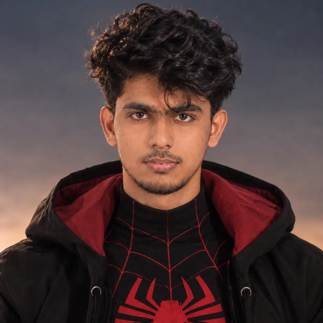 Subject: Front-facing, centered portrait of a young South Asian man with dark, messy, voluminous curly hair and light facial hair/stubble.