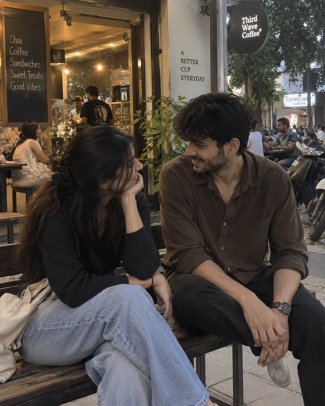 Photorealistic 4:5 vertical candid iPhone photo of a young Indian couple sitting together on a simple bench outside a small local café.
