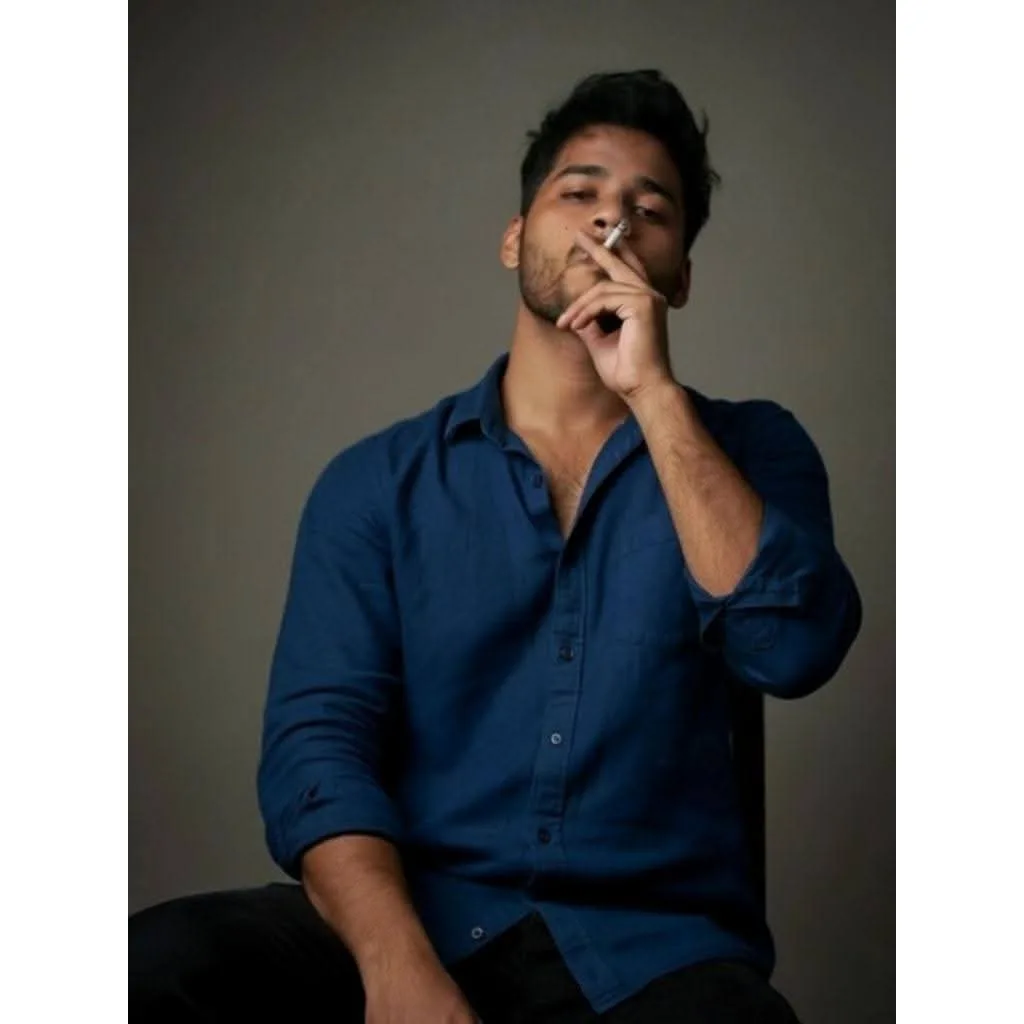 A young South Asian man with dark wavy hair and light facial stubble, sitting and looking slightly upward with a cool, contemplative