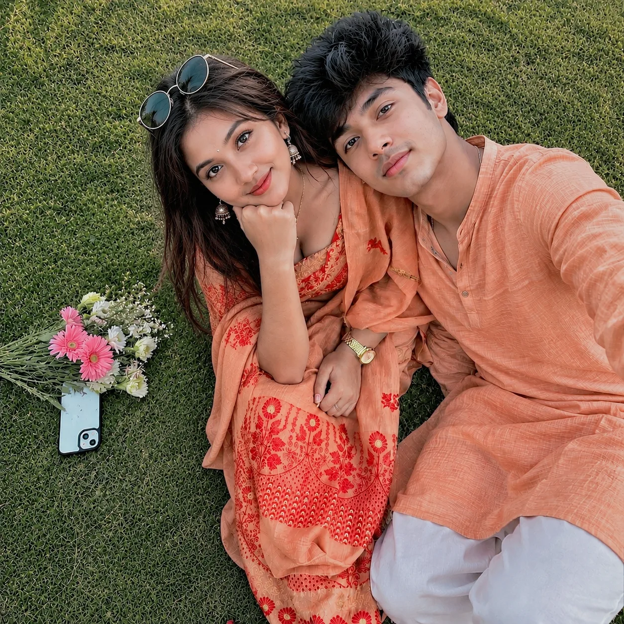 Create a highly realistic, premium smartphone photograph of a young Indian couple lying very close together on a lush green grass lawn,