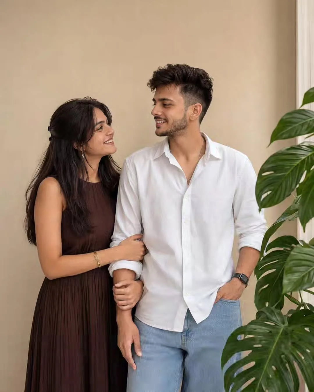 > A high-quality, photorealistic candid portrait of a young South Asian couple standing closely together against a clean warm beige wall in