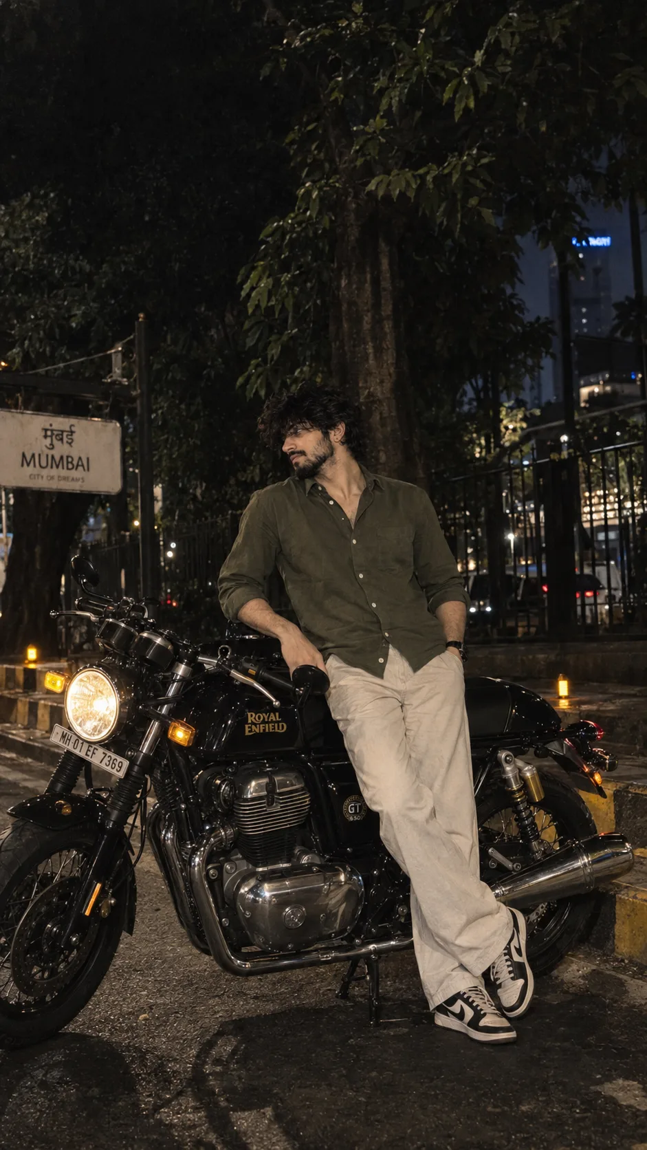A highly detailed, nocturnal photograph capturing a fashionable young man leaning casually against a classic Royal Enfield Continental GT