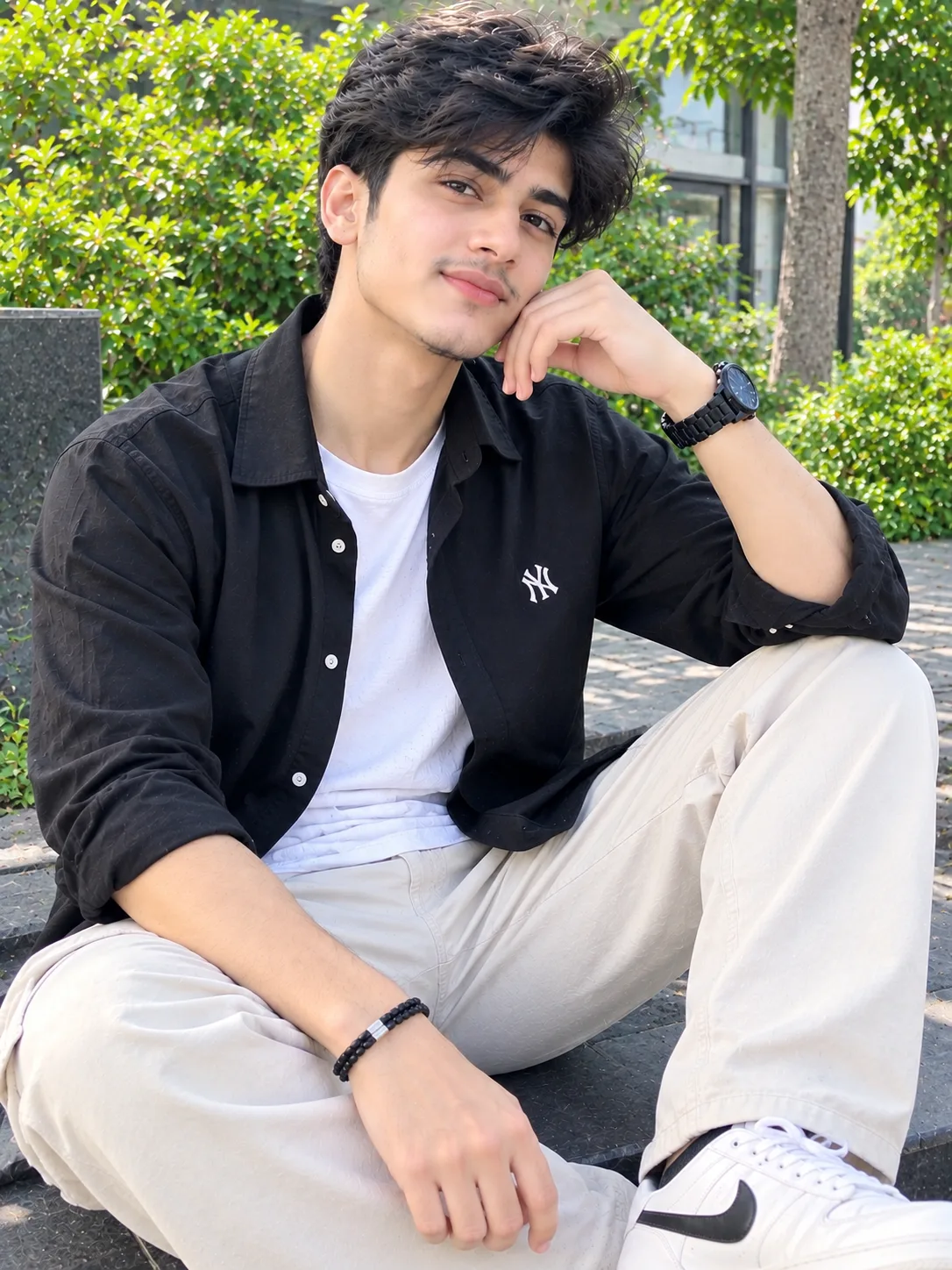A stylish young South Asian man wearing a black open overshirt over a white T-shirt, cream relaxed-fit trousers, and white sneakers.