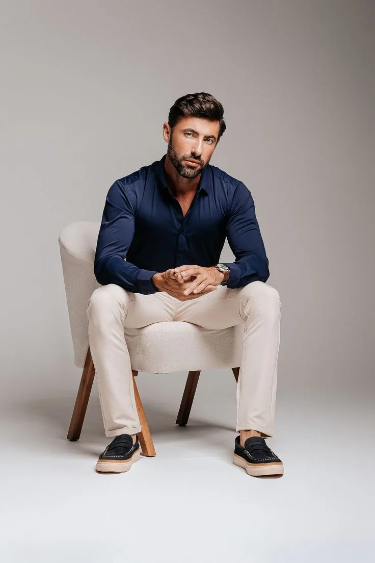 Studio portrait of a confident man sitting on a modern beige armchair with wooden legs, leaning slightly forward with his hands together.