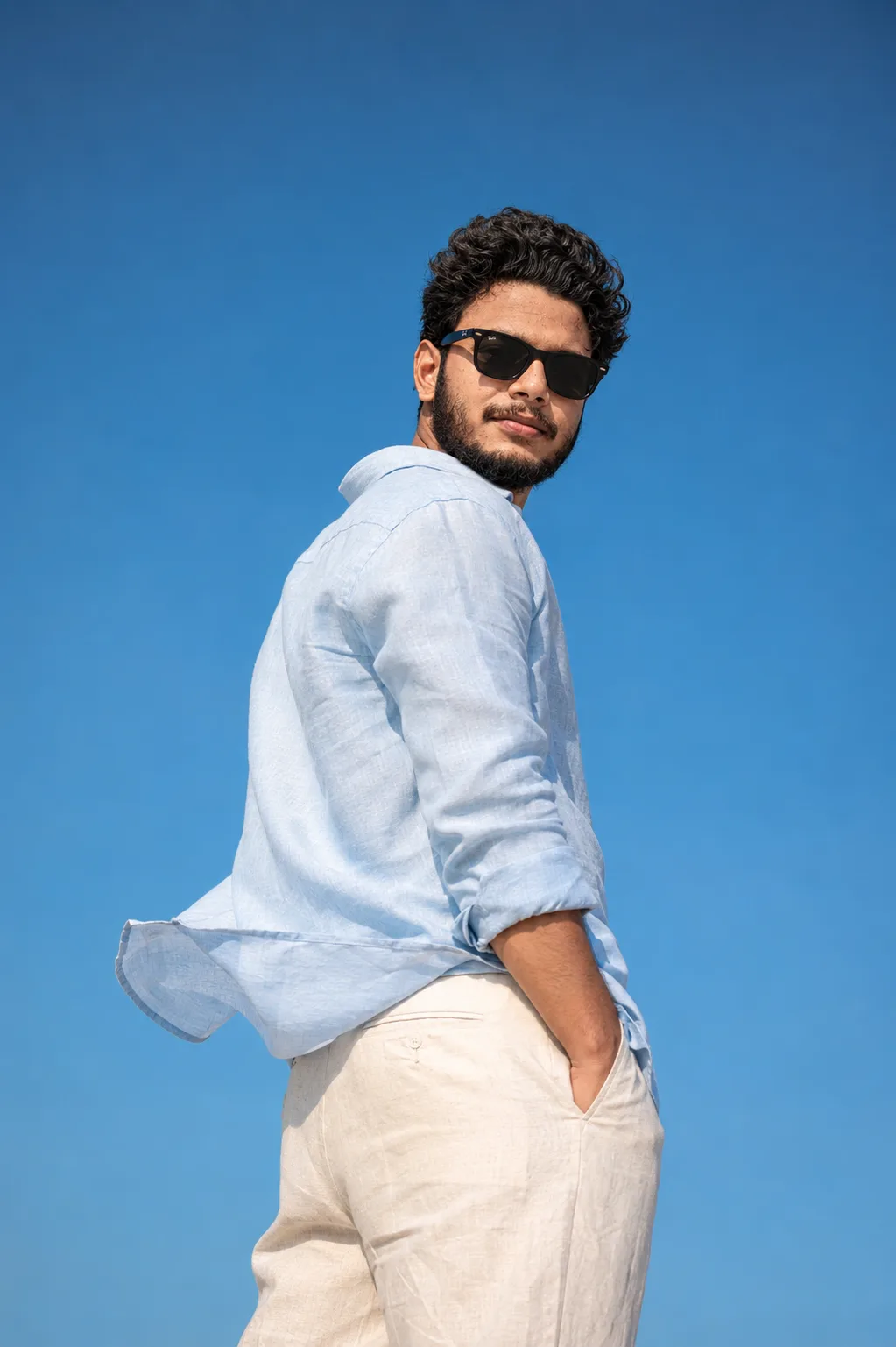 A stylish young man with curly black hair and a neat full beard, looking over his left shoulder towards the camera with a calm, confident