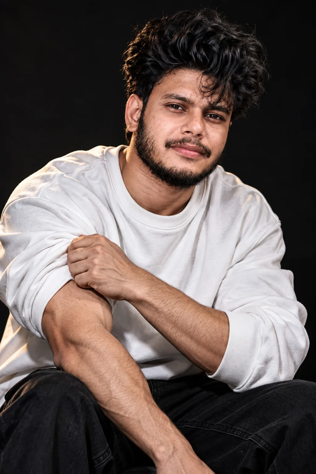 Create an ultra-realistic cinematic studio portrait of a young South Asian/Indian man seated casually against a deep black background.