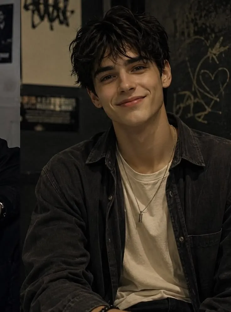 A candid close-up portrait of a handsome young man with messy, textured dark hair, smiling warmly with dimples.