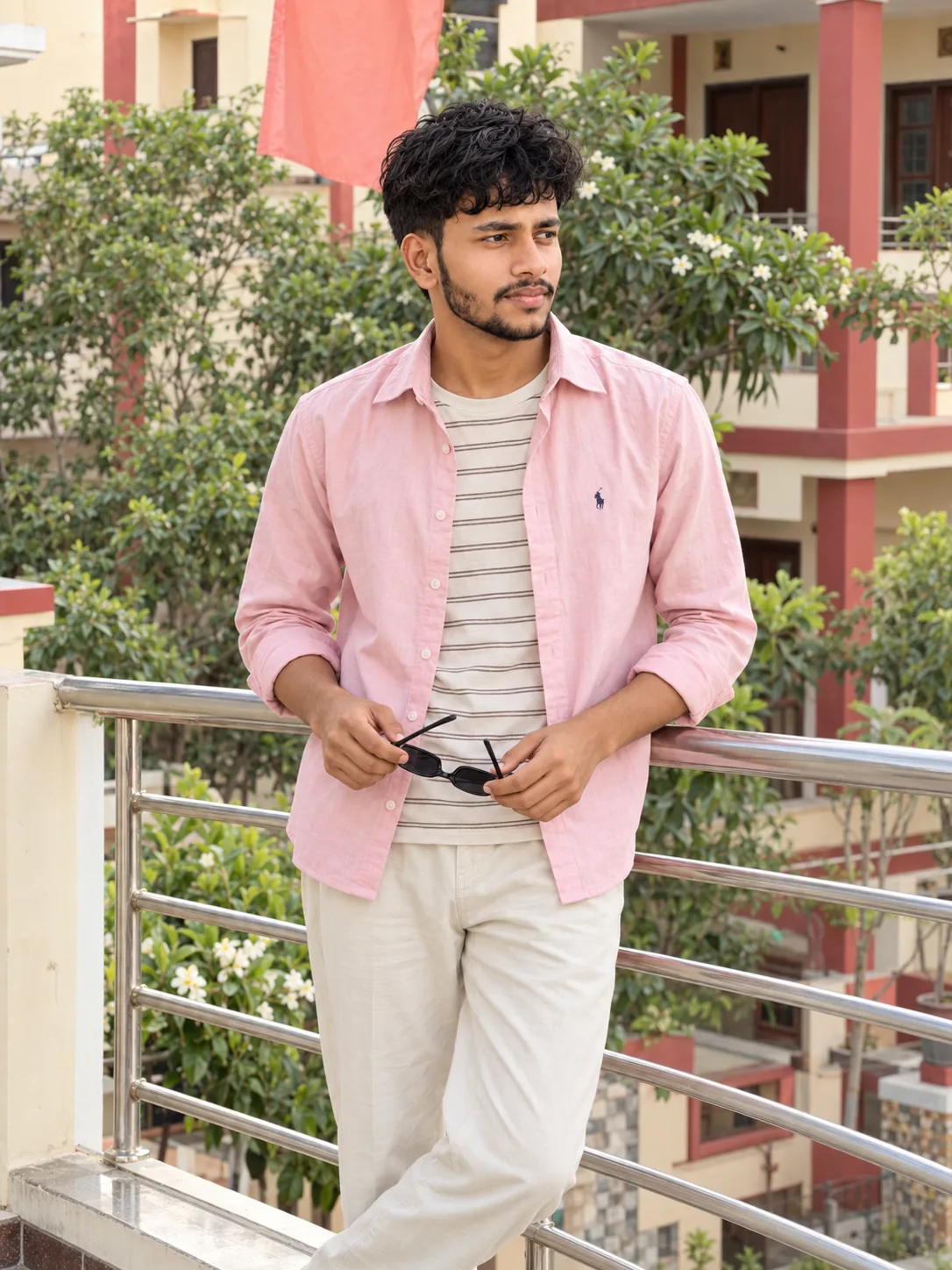 Ultra-realistic professional DSLR photograph of a slim young South Asian man standing casually on an outdoor residential balcony in soft
