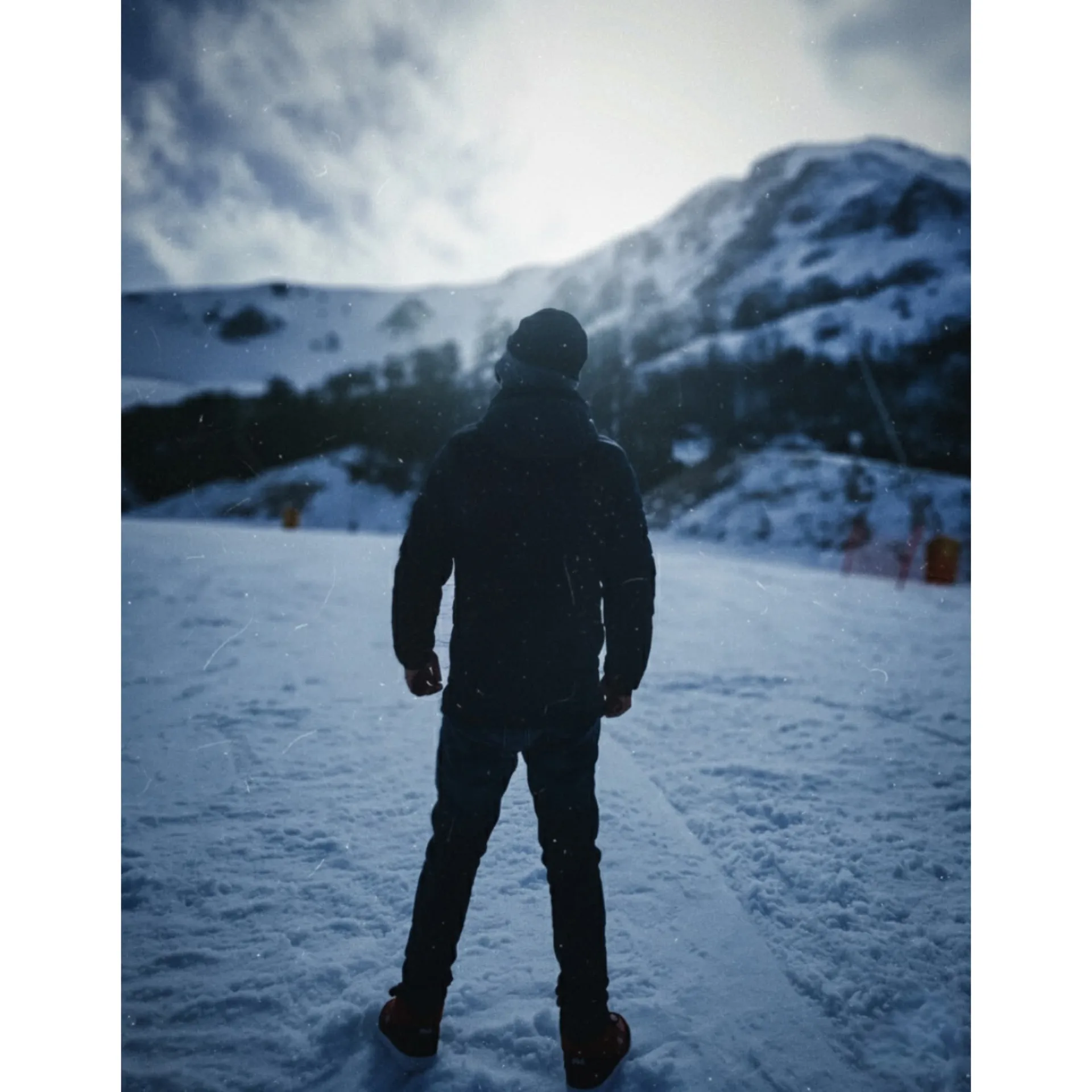 **Prompt:** Cinematic rear view of a lone person standing on a snowy ski slope, facing away from the camera toward distant snow-covered