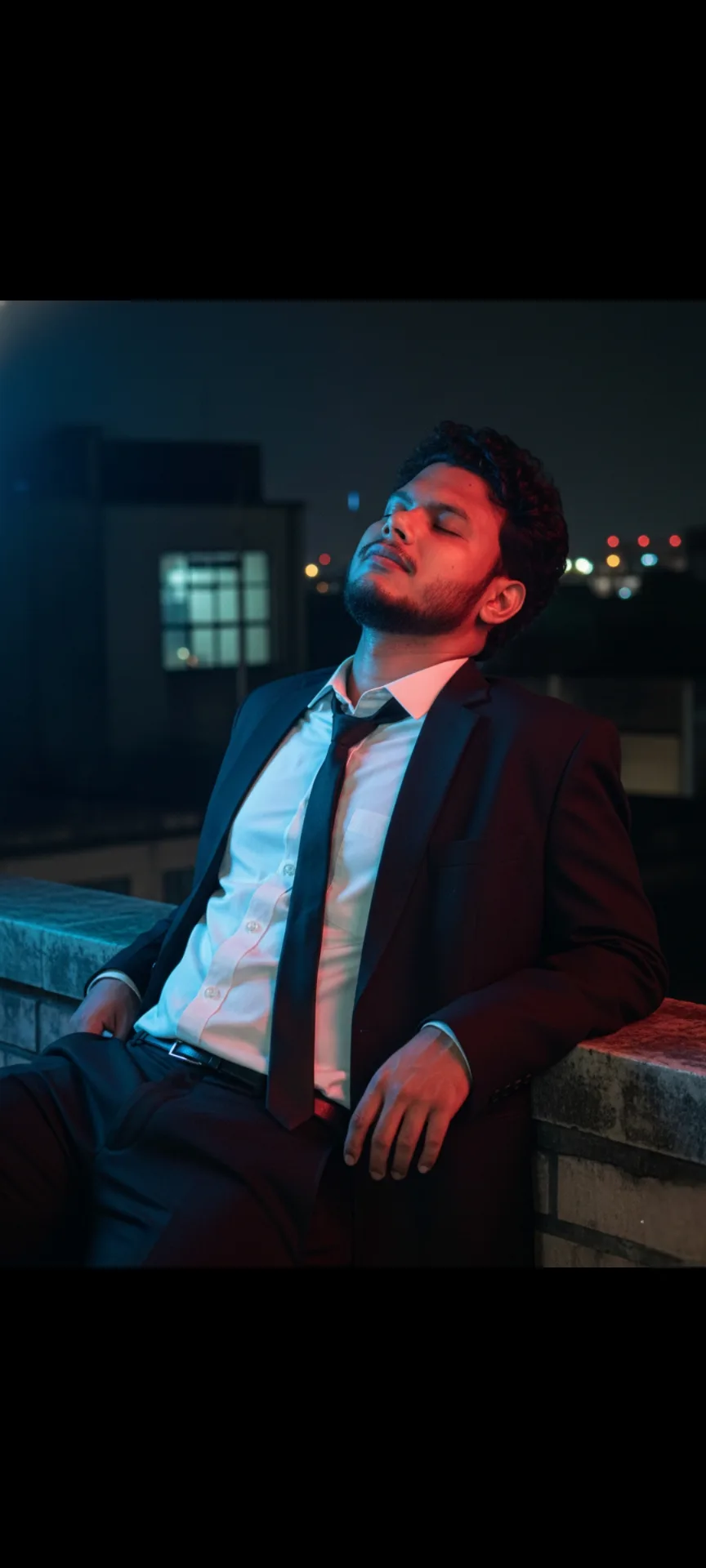 A cinematic, ultra-realistic portrait of a 26-year-old Indian man with a height of 5 feet 5 inches, sitting outdoors on a rooftop at night,