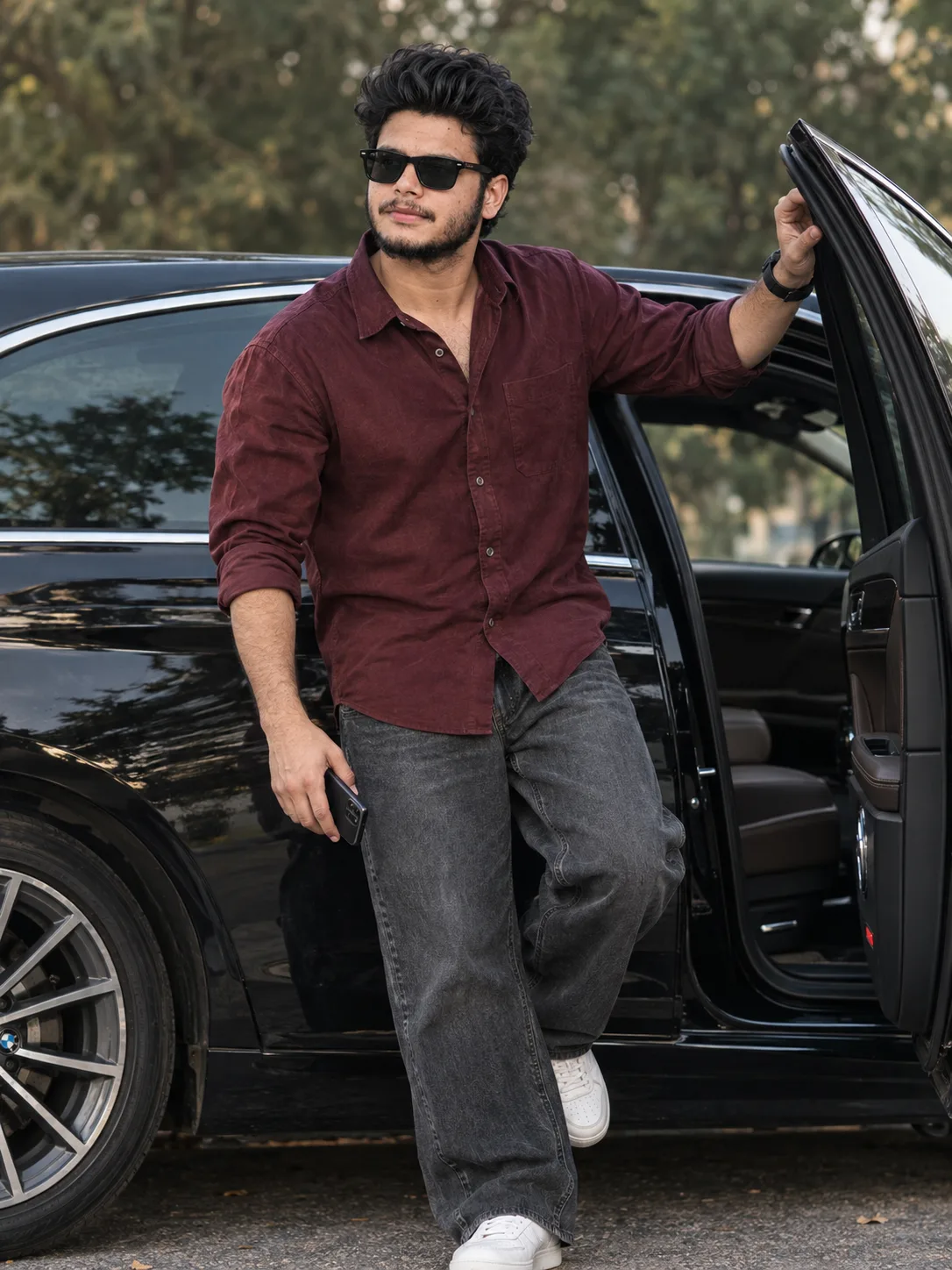 A handsome young South Asian man with curly black hair, full beard, and black rectangular sunglasses stands confidently next to a sleek