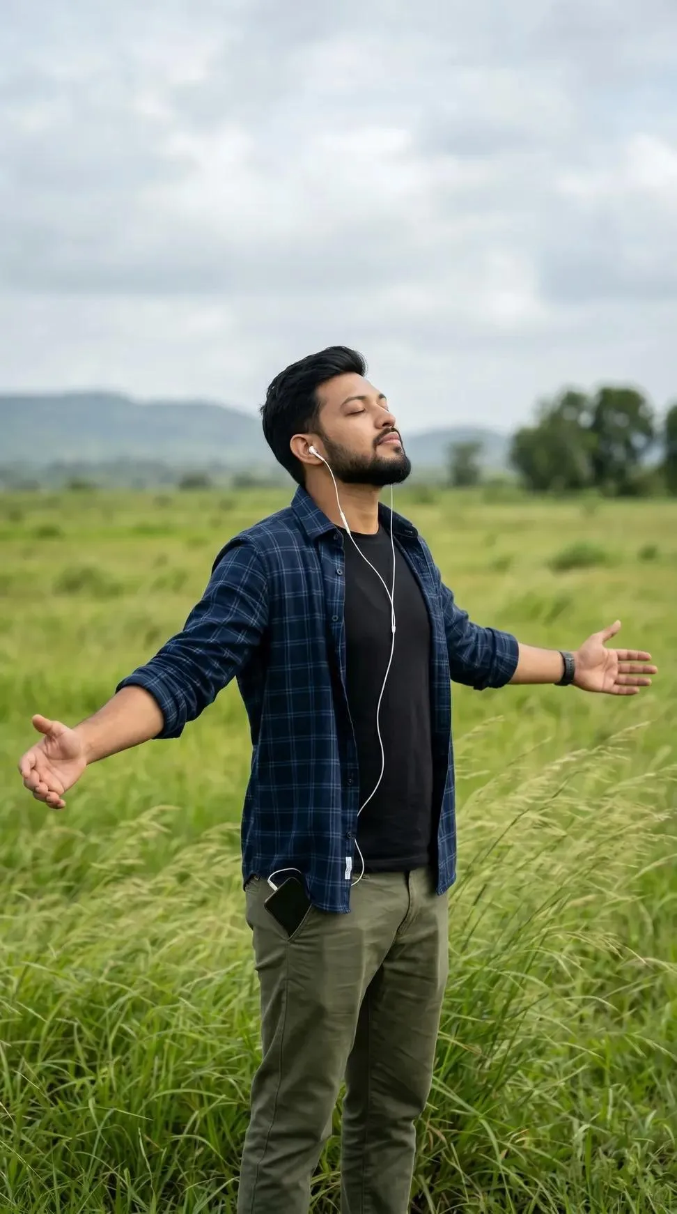 Young handsome Indian man standing in an open green field, eyes closed, face slightly tilted upward, arms spread wide, peaceful vibe.
