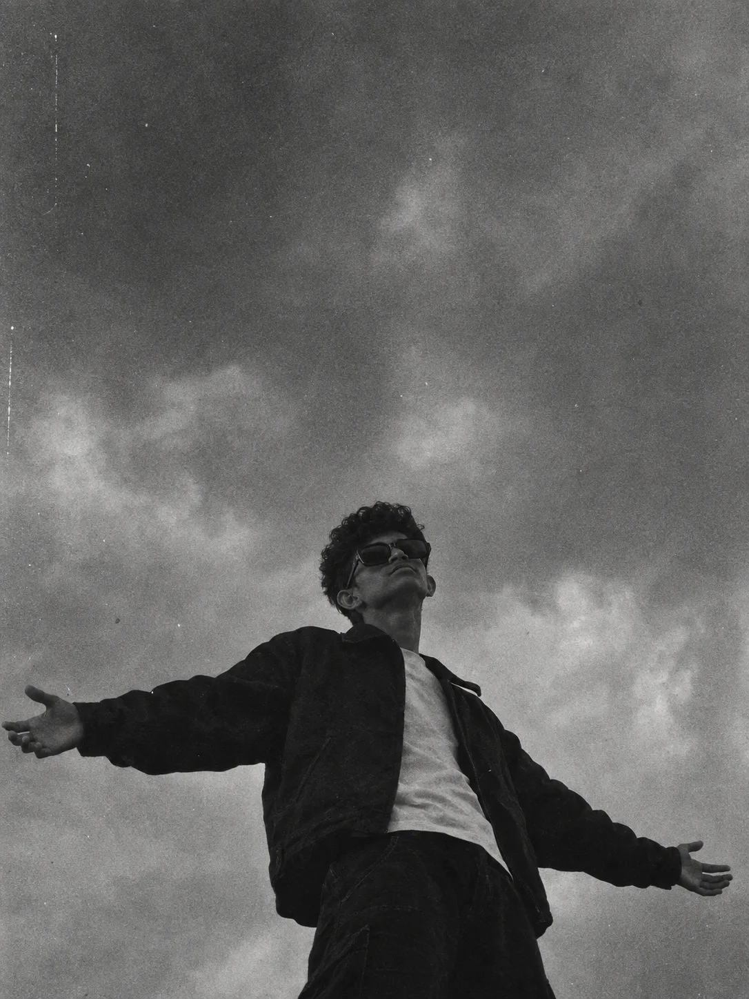 *A black-and-white grainy photo of a person standing with arms outstretched wide against a plain cloudy sky, viewed from a low angle,