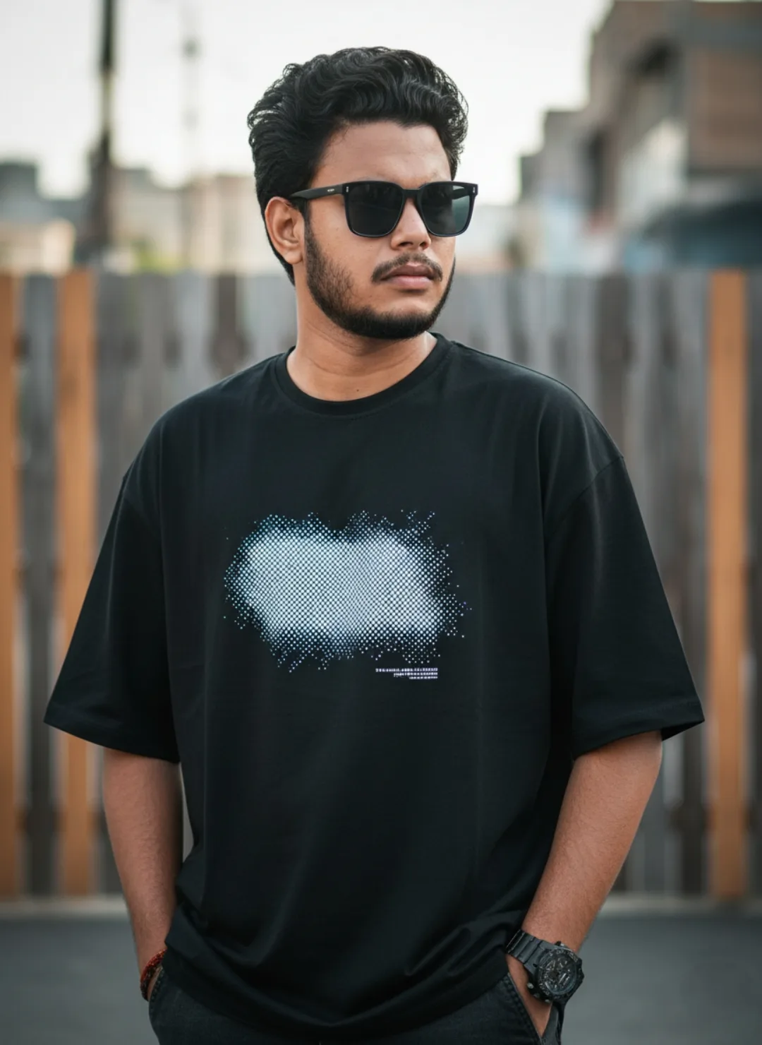 A young South Asian man with short wavy black hair, a neat beard and mustache, wearing black rectangular sunglasses, standing outdoors with