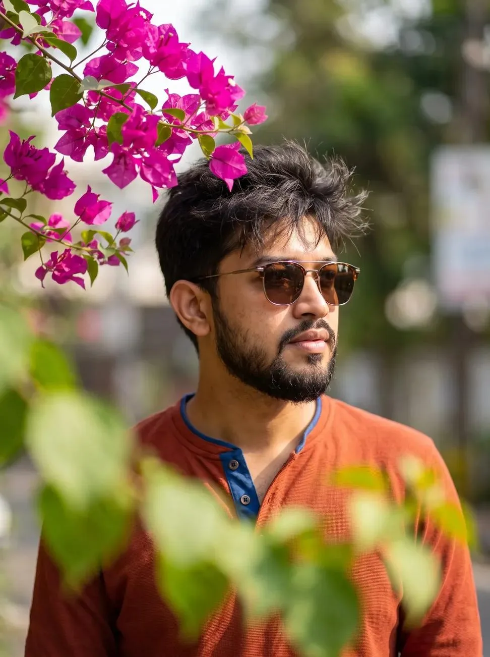 A stylish young man with a slightly messy hairstyle and sunglasses, wearing a vibrant rust-blue Henley shirt.
