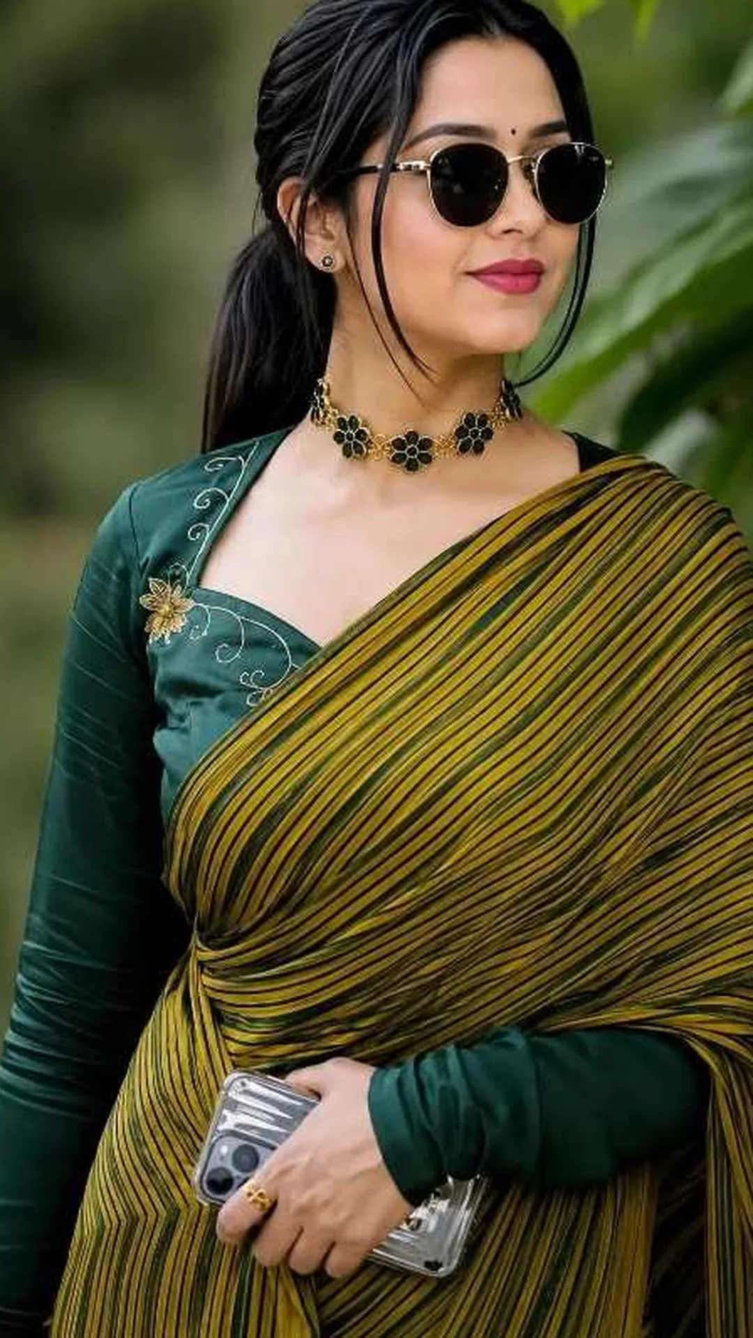 A stylish young Indian woman wearing a dark green blouse with floral embroidery and a yellow and green striped saree, wearing black round