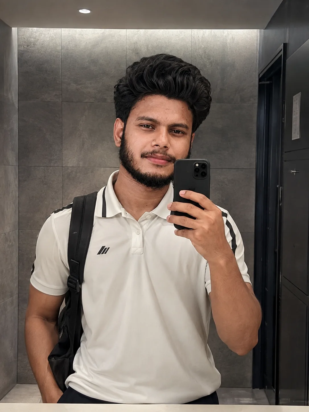 A breathtakingly handsome young male student taking a mirror selfie inside a modern minimalist restroom with large matte gray stone tiles